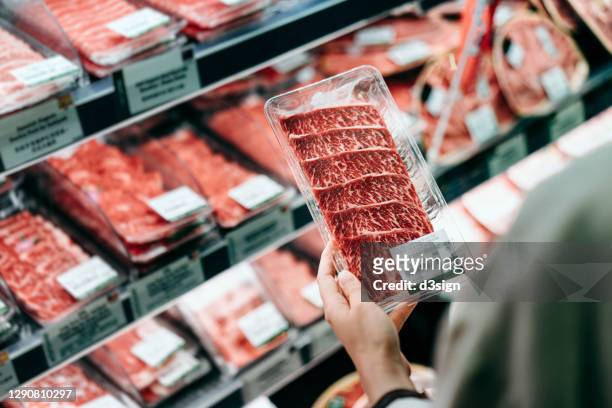 over the shoulder view of young asian woman shopping in a supermarket. she is choosing meat and holding a packet of organic beef in front of the refrigerated section - beef stock pictures, royalty-free photos & images
