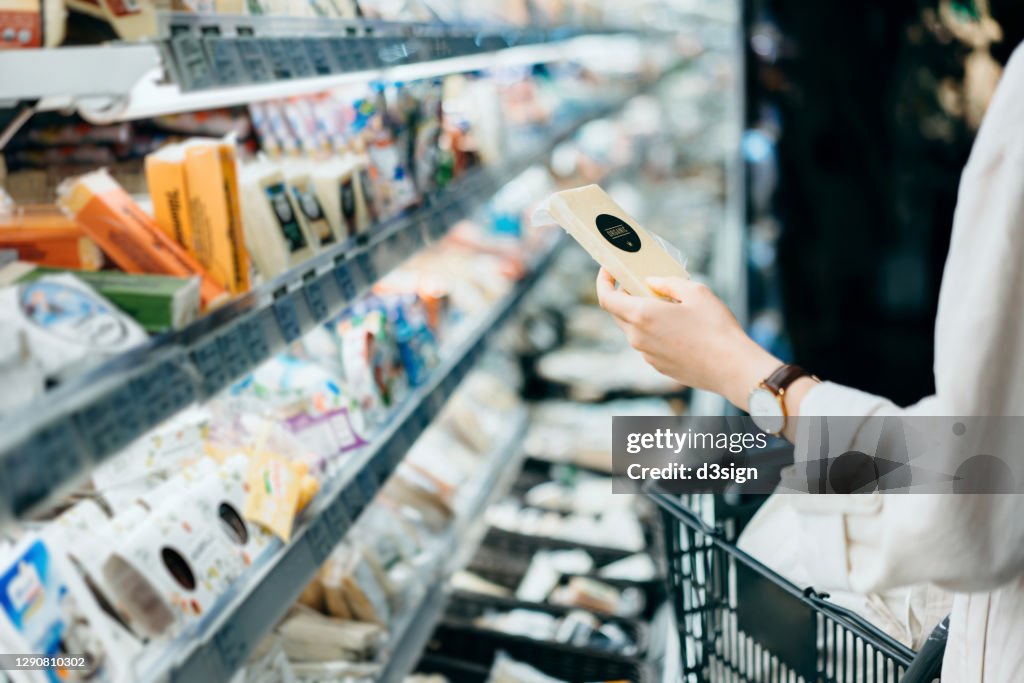 Cropped shot of young Asian woman with a shopping cart, shopping in the dairy section of a supermarket. She is reading the nutrition label on the package of a fresh organic cheese