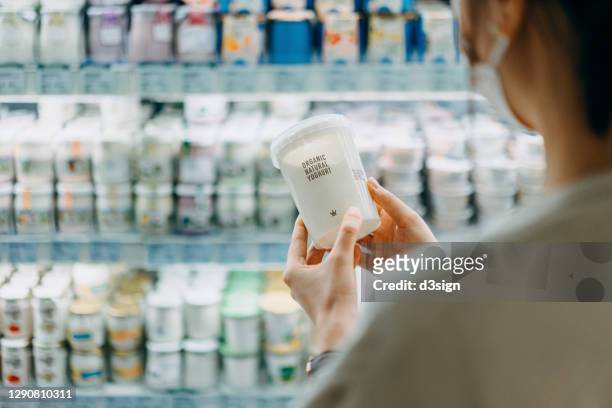 over the shoulder view of young asian woman shopping in the dairy section of a supermarket. she is reading the nutrition label on a container of fresh organic healthy yoghurt - dairy aisle stock pictures, royalty-free photos & images