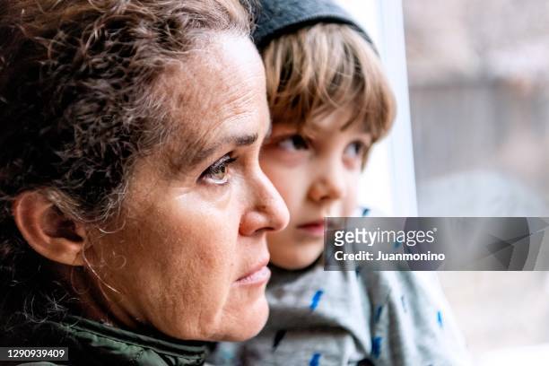 rijpe vrouw die met haar zoon stelt, zeer droevig kijkend door venster dat zich over verlies van haar baan gepast covid-19 pandemie ongerust maakt - pandemic illness stockfoto's en -beelden