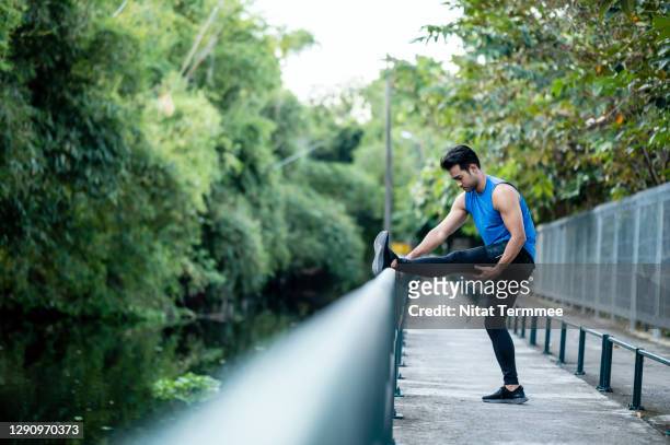 asian sport men workout in next to outside city park, while stretching or warming up his leg on bridge railing. - leg show fotografías e imágenes de stock