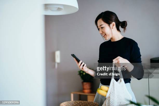 smiling young asian woman coming home from grocery shopping. she is using smartphone while holding a reusable mesh bag full of fresh and healthy organic fruits and vegetables. responsible shopping, zero waste, sustainable lifestyle concept - madre ama de casa fotografías e imágenes de stock