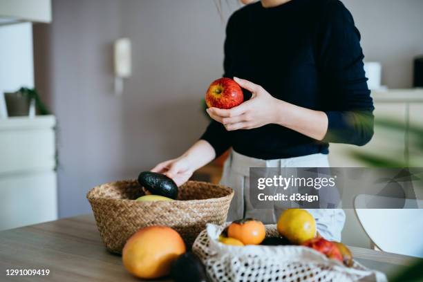 cropped shot of young asian woman coming home from grocery shopping, unpacking and organizing fresh and healthy organic fruits and veggies from a reusable bag to a rattan basket on the table. responsible shopping, zero waste, sustainable lifestyle concept - fair trade stock-fotos und bilder