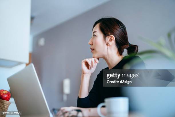 young asian woman in deep thought and looking away while working from home with laptop - deep conversation stock pictures, royalty-free photos & images