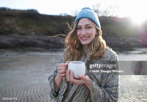 woman wearing bobble hat on winter beach with hot drink. - bobble hat stock pictures, royalty-free photos & images