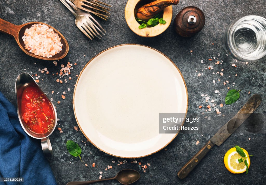 Empty plate and kitchen utensils and ingredients over dark background.