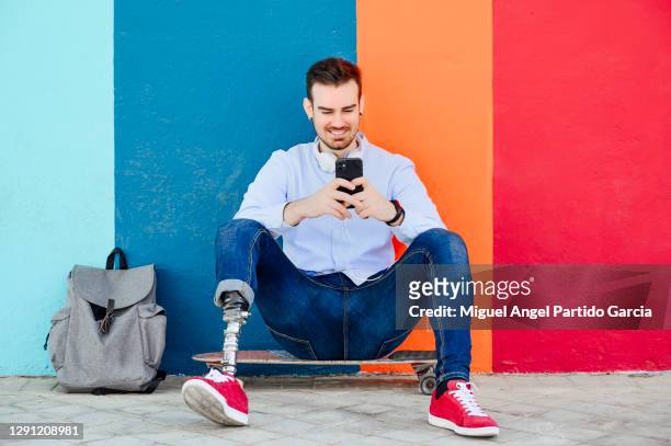 Smiling young man with leg prosthesis and skateboard using smartphone