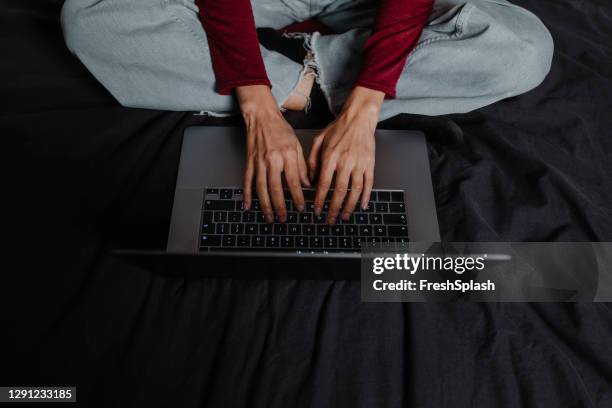 hands of an anonymous woman typing on a laptop keyboard while sitting cross-legged on her bed - dark humor stock pictures, royalty-free photos & images