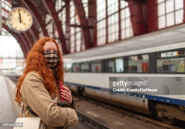 student girl wearing a face mask stands at the tracks on the railway platform with clock behind her - belgium stock pictures, royalty-free photos & images