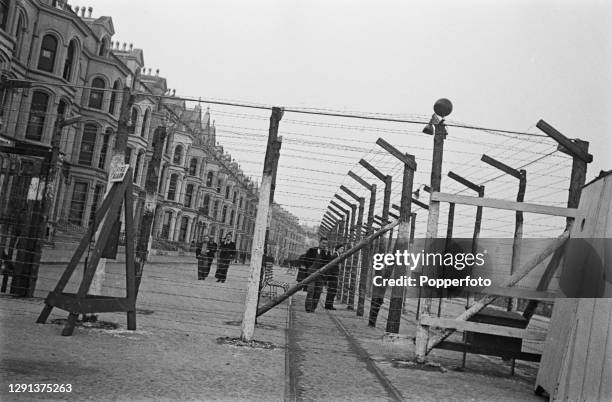 Group of internees walk alongside barbed wire barricades outside their Internment Camp, comprising former hotels and boarding houses, overlooking...