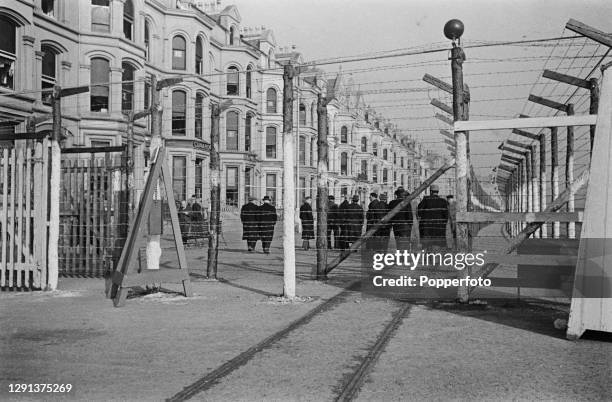 Group of internees walk alongside barbed wire barricades outside their Internment Camp, comprising former hotels and boarding houses, overlooking...