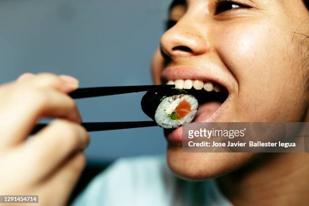 close-up of a young girl taking a delicious piece of sushi, maki, to her mouth with her chopsticks. neutral background - stäbchen stock-fotos und bilder