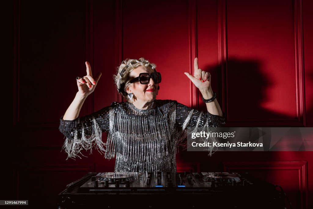 Old woman stands with headphones at the dj console and plays music and dances
