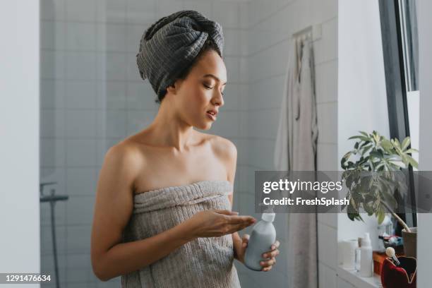 joven mujer afroamericana envuelta en una toalla después de una ducha sosteniendo un producto para el cuidado de la piel - shampoo fotografías e imágenes de stock