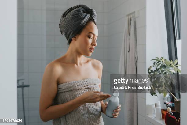 young african american woman wrapped in a towel after a shower holding a skin care product - hair conditioner stock pictures, royalty-free photos & images