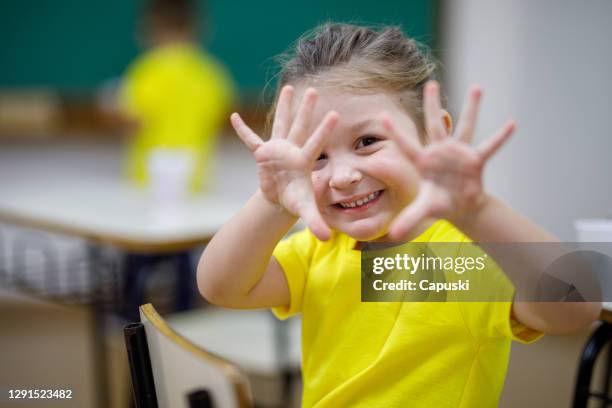 lächelndes kleines mädchen in einem klassenzimmer zeigt seine hände und blicke in die kamera - raised finger stock-fotos und bilder