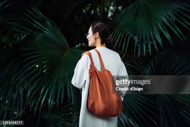 rear view of smiling young asian woman relaxing in a park. she is carrying a brown reusable bag against green nature plants. responsible shopping, zero waste and sustainable lifestyle concept - トートバッグ ストックフォトと画像