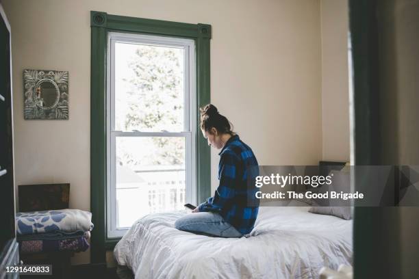 young woman sitting on bed looking at smart phone - brunette woman bed stockfoto's en -beelden