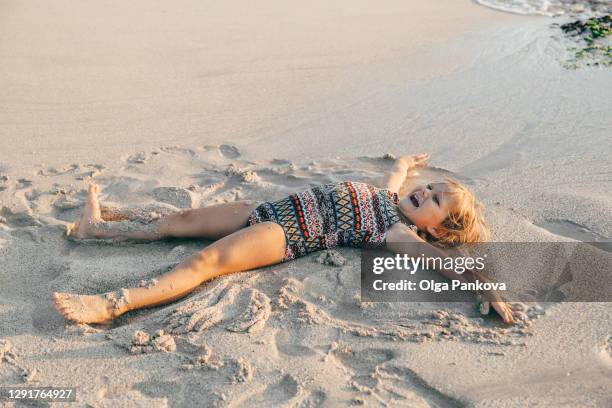 toddler girl having fun in the sand on the beach during sunset. playfully creating a sand angel. the indian ocean coast. bali island. beautiful view. family leisure. - angel island imagens e fotografias de stock
