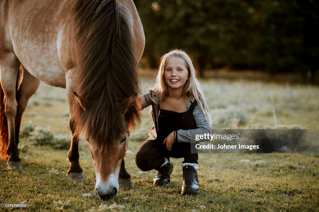 Smiling girl crouching on meadow with grazing pony