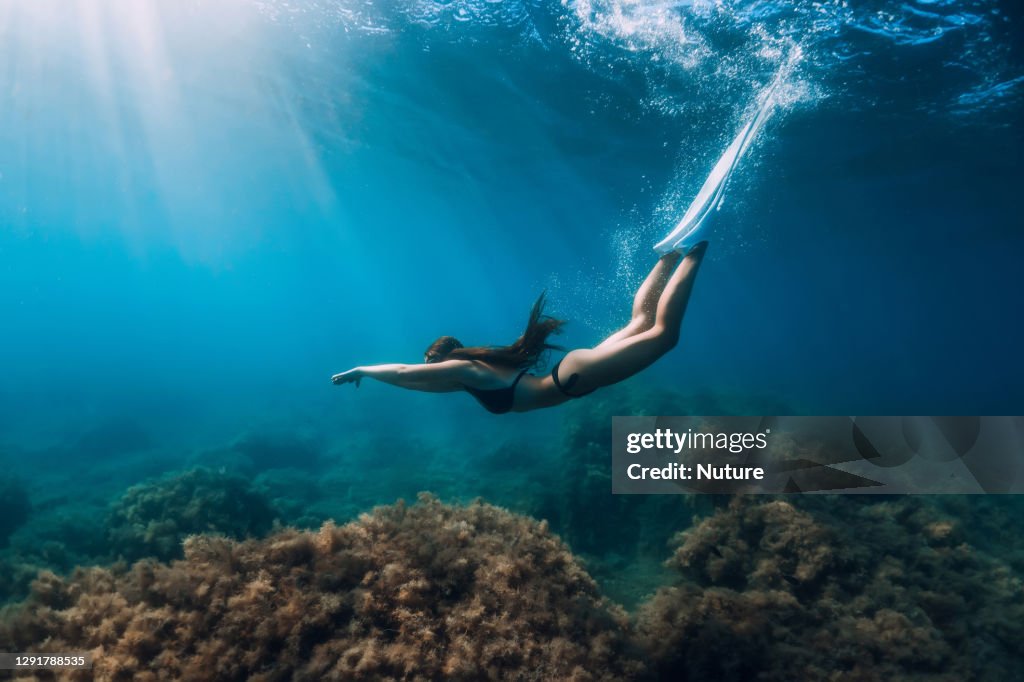 Freediver girl with white fins glides underwater with amazing sun rays and seaweed. Freediving underwater in blue sea