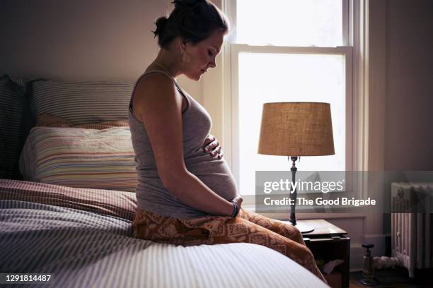 pregnant young woman sitting on bed at home - pregnant woman stock pictures, royalty-free photos & images