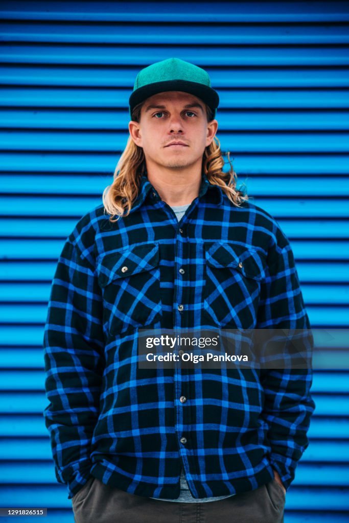 A long hair man in a green cap and a blue plaid jacket against a blue wall.