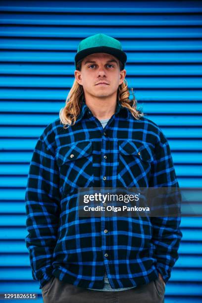 a long hair man in a green cap and a blue plaid jacket against a blue wall. - monocromo vestuário - fotografias e filmes do acervo