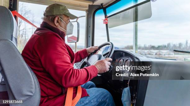 senior man, the bus driver wearing the protective n95 mask because of the covid-19 pandemic, driving a school bus in the rural area. - alex potemkin coronavirus stock pictures, royalty-free photos & images