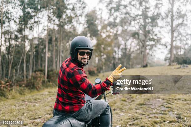 smiling male biker gesturing while exploring forest on motorcycle - victory sign man stockfoto's en -beelden