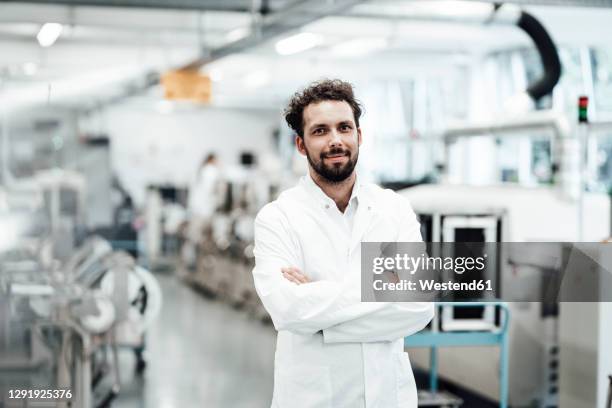 confident male scientist in white lab coat while standing with arms crossed at bright laboratory - industrie und mensch stock-fotos und bilder