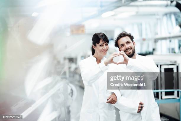 smiling male and female scientists making heart shape with hands while standing arm in arm at laboratory - scientist standing stock-fotos und bilder