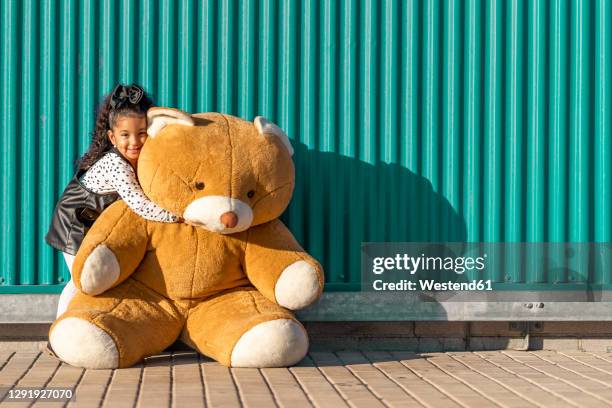 cute girl embracing teddy bear while standing against green wall - teddy bear stock pictures, royalty-free photos & images
