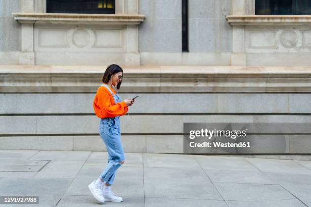 young woman using mobile phone while walking against wall on footpath - phone side view stock pictures, royalty-free photos & images