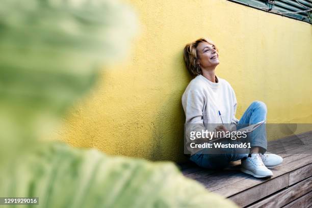 thoughtful smiling woman sitting with diary against yellow wall on building terrace - 2020 diary stock pictures, royalty-free photos & images