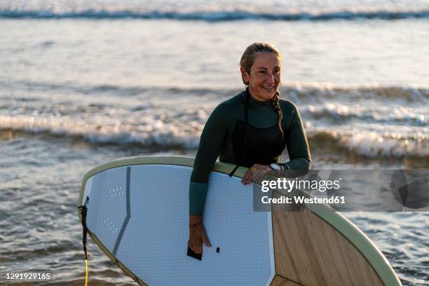 smiling woman holding paddleboard standing in sea at dawn - paddleboard stock-fotos und bilder