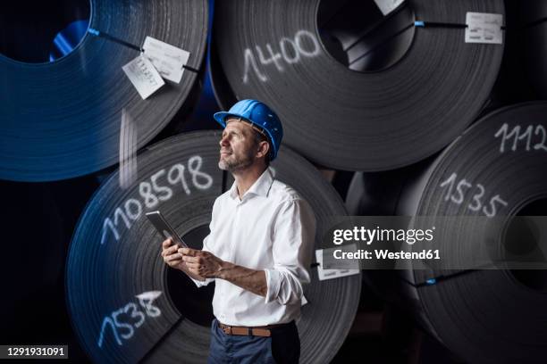 businessman holding digital tablet while standing by steel rolls at industry - metallindustrie stock-fotos und bilder