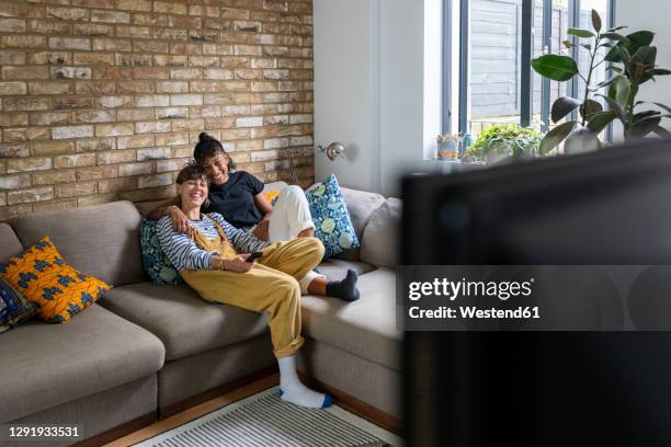 smiling lesbian couple watching television while sitting on sofa at home - couple tv stock pictures, royalty-free photos & images