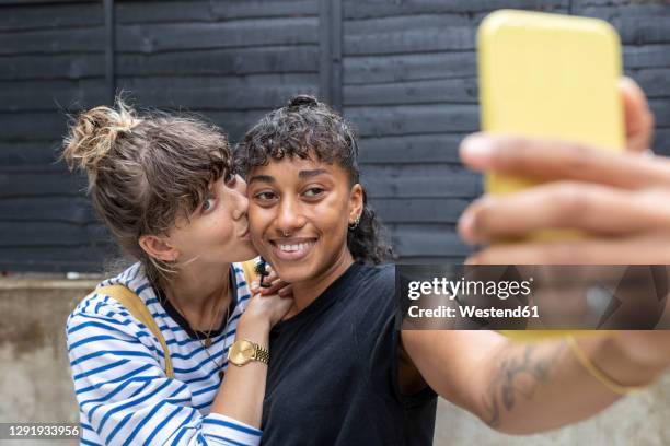 smiling woman taking selfie on mobile phone while girlfriend kissing her standing at back yard - photos of lesbians kissing stock pictures, royalty-free photos & images