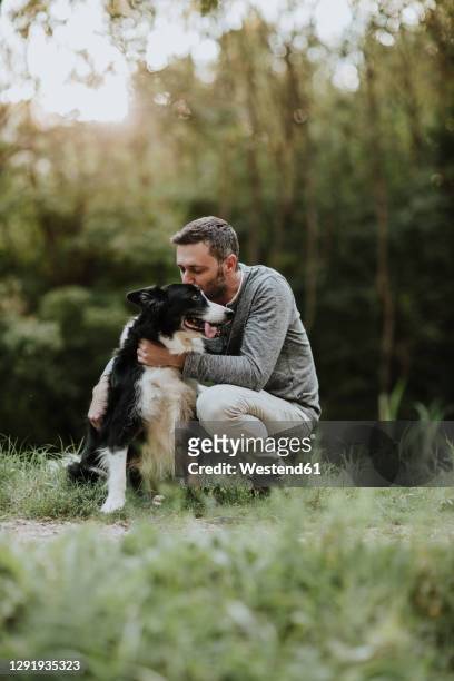 man kissing his dog while crouching at park - chien et maitre photos et images de collection
