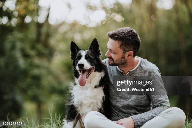 smiling man staring dog while sitting at public park - huisdiereigenaar stockfoto's en -beelden