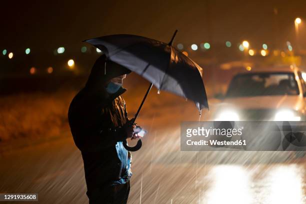 young man with umbrella and face mask using mobile phone while standing against breakdown car during rainfall - stranded 個照片及圖片檔