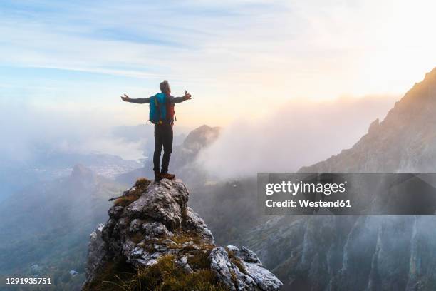 pensive hiker with arms outstretched standing on mountain peak during sunrise at bergamasque alps, italy - arms outstretched fotografías e imágenes de stock