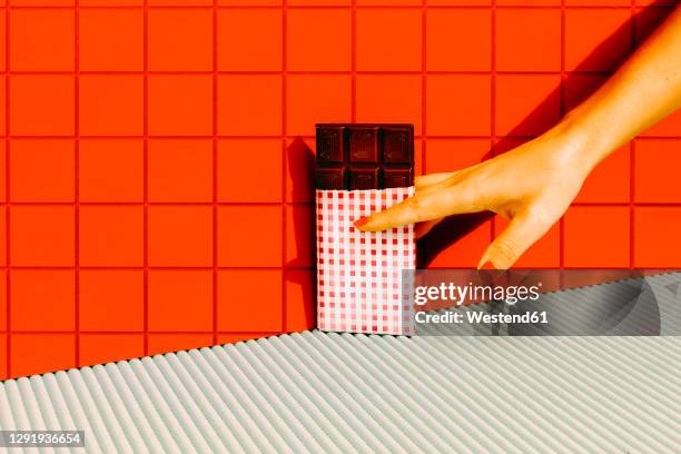 woman hand holding dark chocolate bar against red tile wall - chocolat noir photos et images de collection