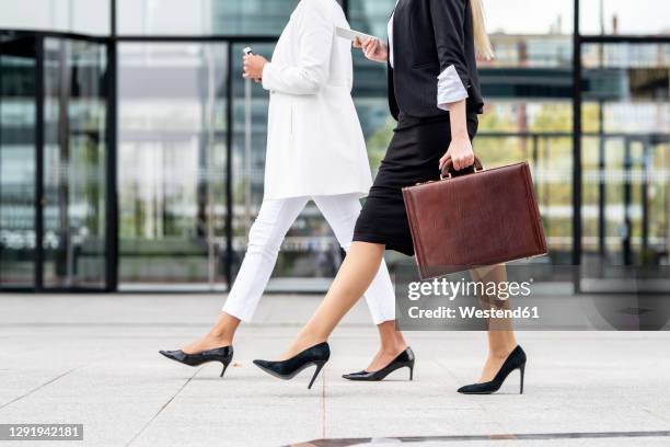 businesswoman with briefcase using mobile phone while walking by colleague on footpath - attaché stockfoto's en -beelden