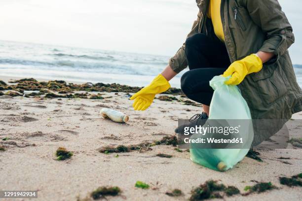female environmentalist collecting bottle in garbage bag while crouching at beach - volunteer beach photos et images de collection