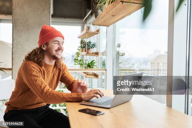 smiling man wearing knit hat using laptop sitting at home - brown hat ストックフォトと画像