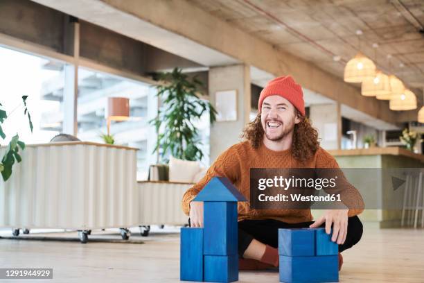 cheerful man playing with toy block while sitting on floor in living room at home - brown hair ストックフォトと画像