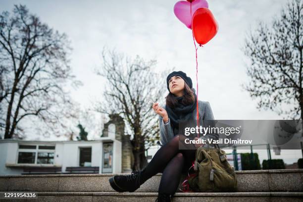 young attractive woman sitting and smoking with heart - shaped balloons in her hand. being late for a date / love disappointment / lonely on a saint valentine's day concept. - saint valentin stock pictures, royalty-free photos & images