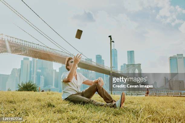 young man sitting outdoors in park throwing phone mid air. - throwing phone stock pictures, royalty-free photos & images